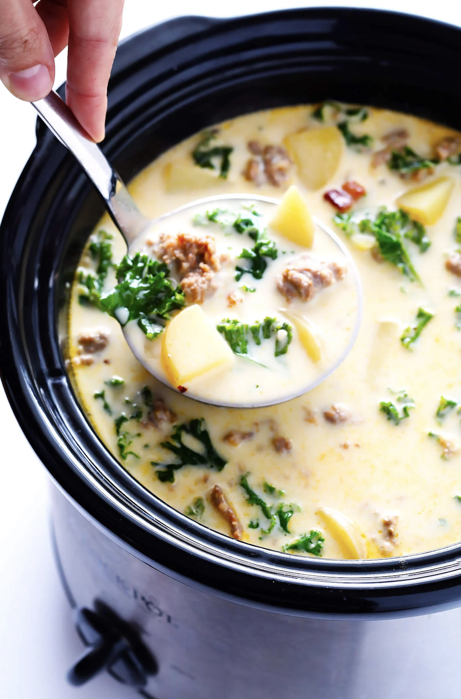 Hand serving soup from a crockpot with sausage, potatoes, and kale