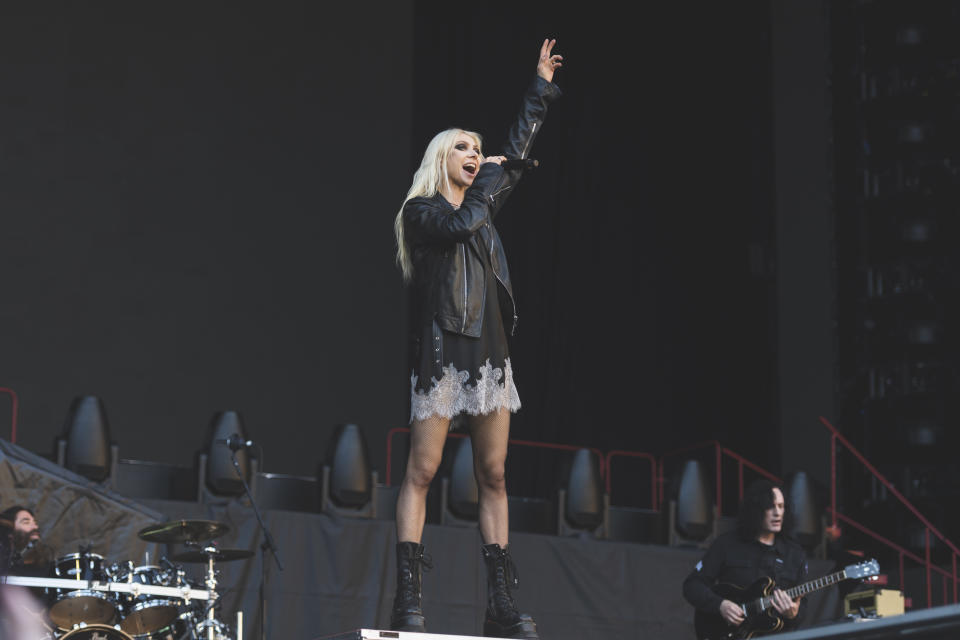 Taylor Momsen performing on stage, wearing a black leather jacket, lace-trimmed dress, and black boots, with band members playing instruments in the background