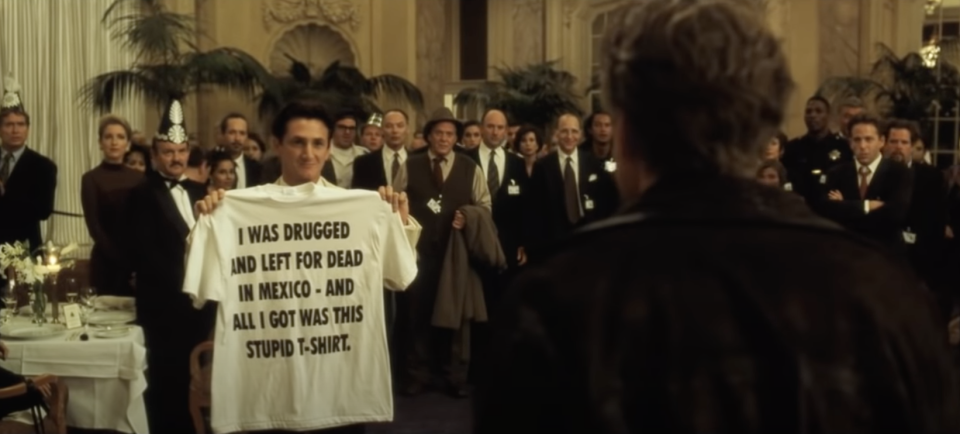 A man at a party holding up a shirt that reads: "I was drugged and left for dead in Mexico, and all I got was this stupid t-shirt"