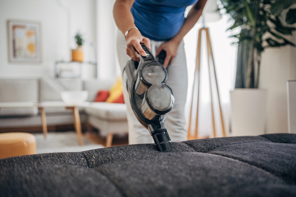 A person uses a handheld vacuum cleaner to clean a couch in a living room. The image focuses on the action of vacuuming