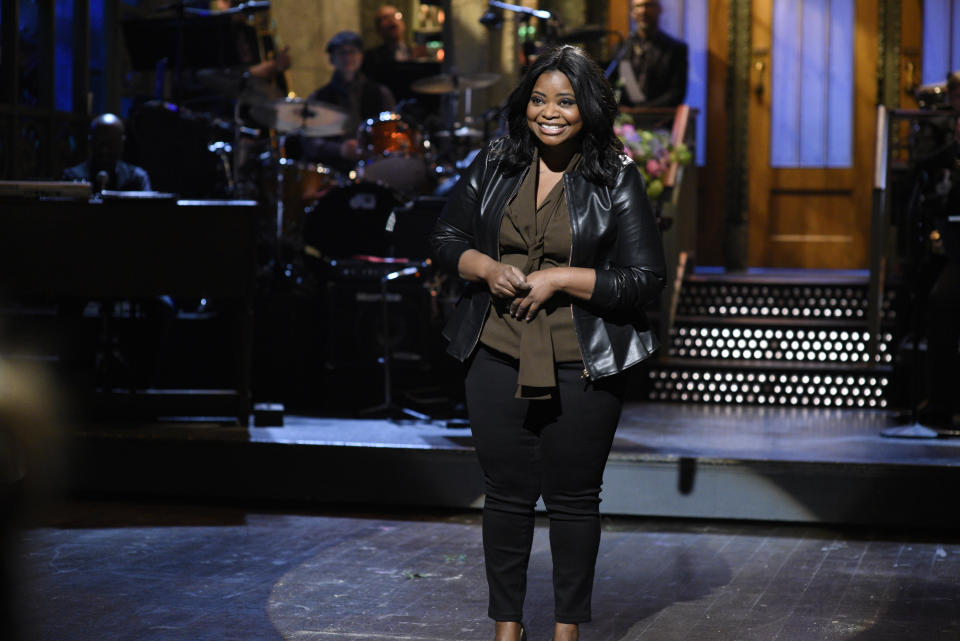 Woman smiles on stage with microphone, band in background, wearing a leather jacket and black pants