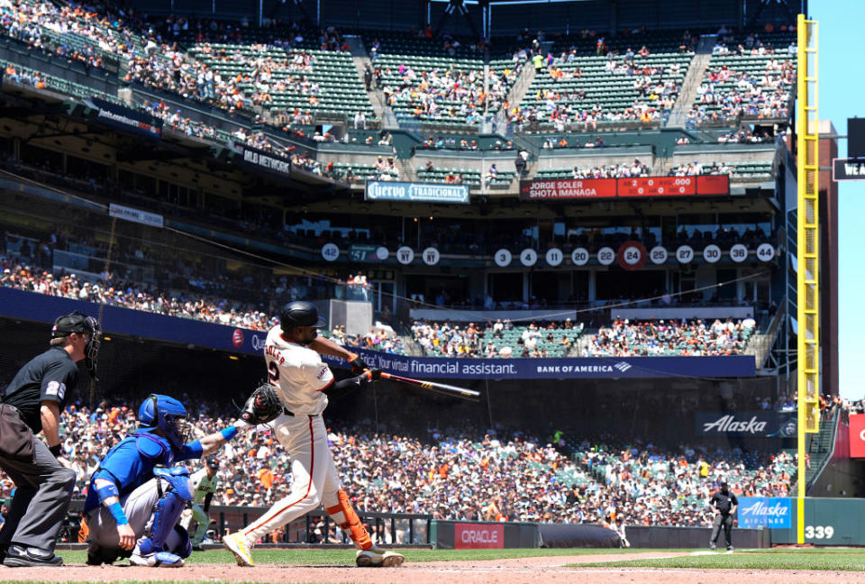 A baseball player from the San Francisco Giants is hitting a ball during a game at a stadium with a large crowd. An umpire and catcher are also visible