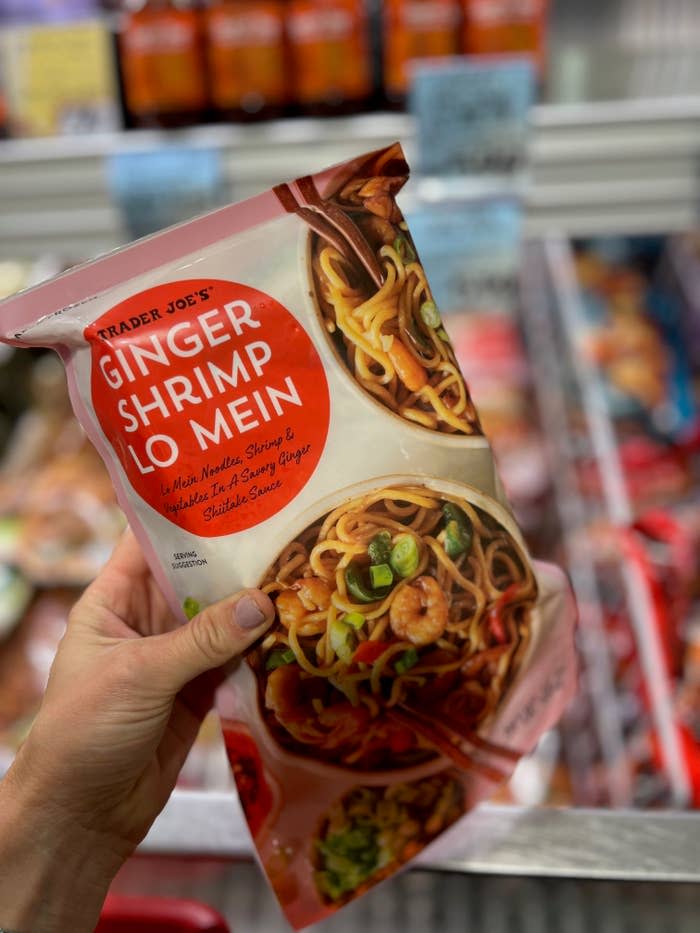 A hand holding a package of Trader Joe's Ginger Shrimp Lo Mein in a grocery store freezer section. The package features noodles, shrimp, and vegetables