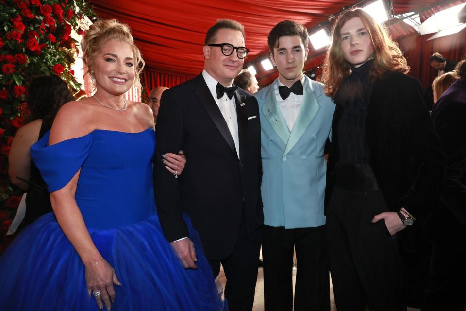 jeanne moore, brendan fraser, holden fraser, and leland fraser smile for a photo together while standing inside a red tent, moore wears a royal blue gown, brendan wears a black tuxedo, holden wears a baby blue suit jacket with a black bowtie and white shirt, leland wears an all black suit