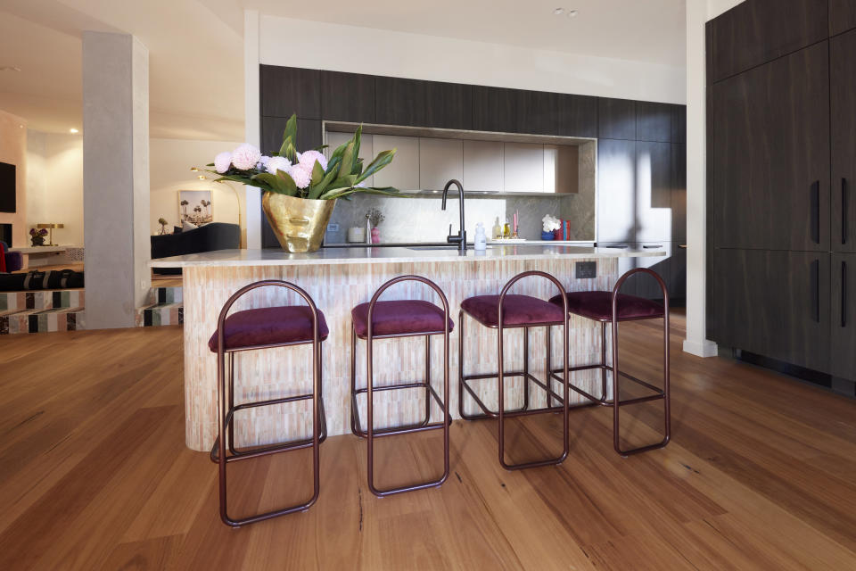 A shot of the kitchen with four purple coloured stool in front, a large vase on the bench