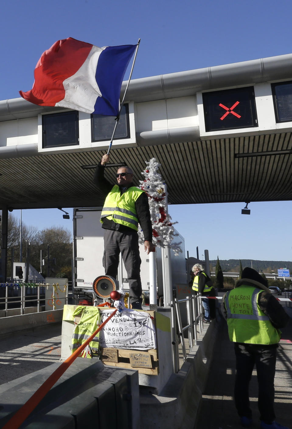 A demonstrators wearing a yellow vest waves a French flag as demonstrators open the toll gates on a motorway near Aix-en-Provence, southeastern France, Tuesday, Dec. 4, 2018. French Prime Minister Edouard Philippe announced a suspension of fuel tax hikes Tuesday, a major U-turn in an effort to appease a protest movement that has radicalized and plunged Paris into chaos last weekend. (AP Photo/Claude Paris)