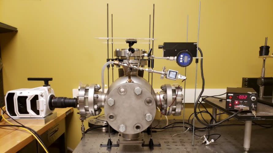  A table-sized metal chamber sealed with four large bolts, connected to other mechanical devices with tubes and wires across three different table surfaces in a room with yellow walls. 