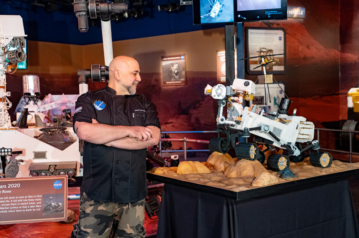  A man in a black polo shirt stands in front of a life-size model of nasa's perseverance mars rover. 