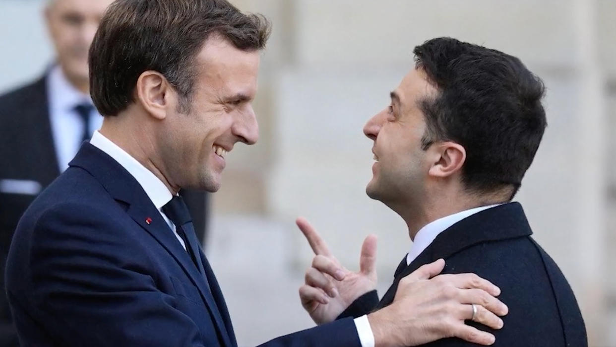 French President Emmanuel Macron, smiling broadly, puts a reassuring hand on the shoulder of Ukrainian President Volodymyr Zelensky, who points upward with great enthusiasm.