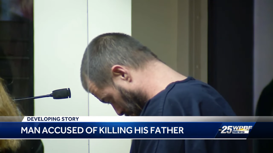 Man with head bowed in blue attire before a microphone, news caption about a developing story on patricide