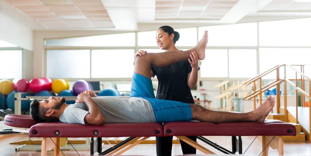 male patient lying on massage table with leg held by massage therapist, fascia stretch therapy