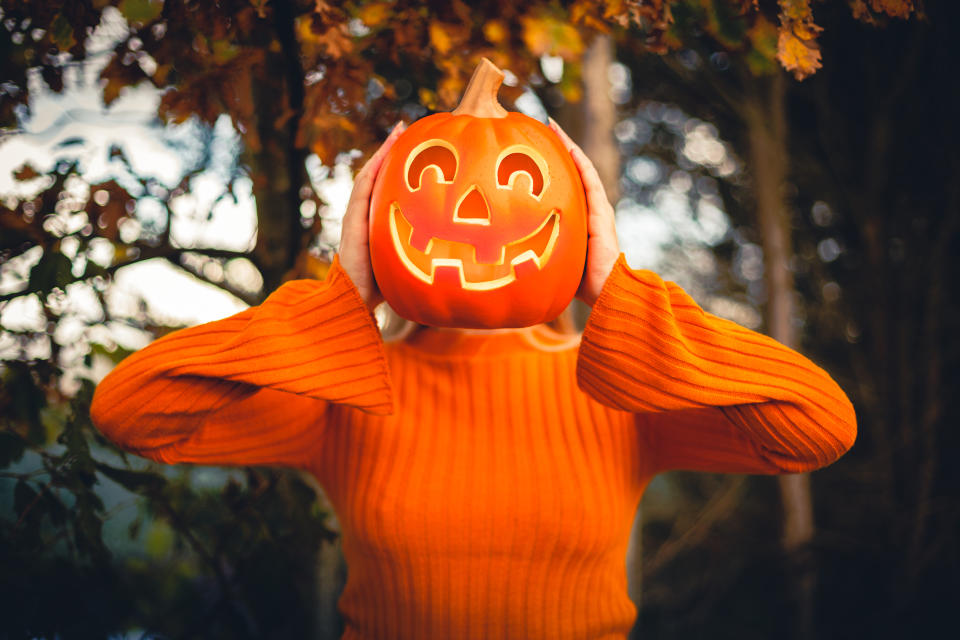 Happy young woman who is in orange jumper, holding a halloween pumpkin against her face at outside