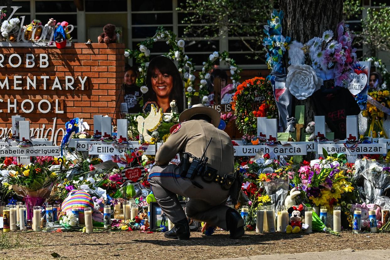 A uniformed police officer crouches in front of a memorial made from white crosses, candles and flowers around the sign in front of Robb Elementary School.