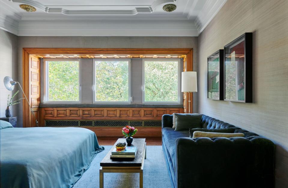 a king bed has a blue linen cover, a dark blue velvet sofa against a wall covered in a sea grass wallpaper, a floor lamp, a brass and slate cocktail table, and carved woodworking around three windows