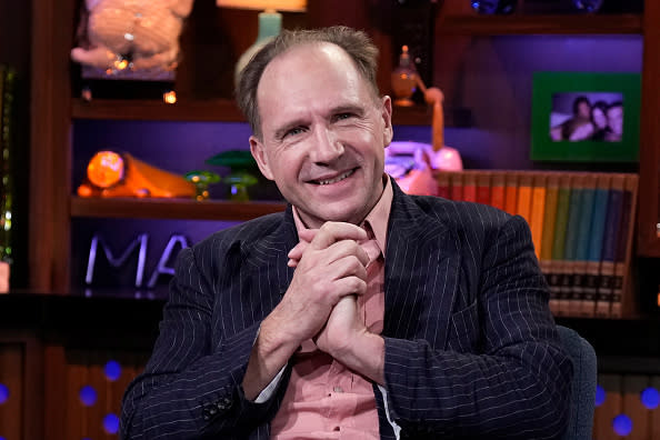 Ralph Fiennes smiling and clasping his hands while sitting during an interview. He is wearing a pinstripe blazer and a collared shirt underneath