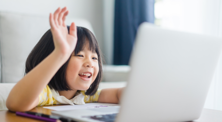 Image of a young girl raising her hand in front of a laptop.