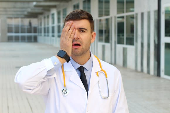 Doctor standing in a hallway and wearing a stethoscope, covering his right eye with his right hand, with his mouth agape.