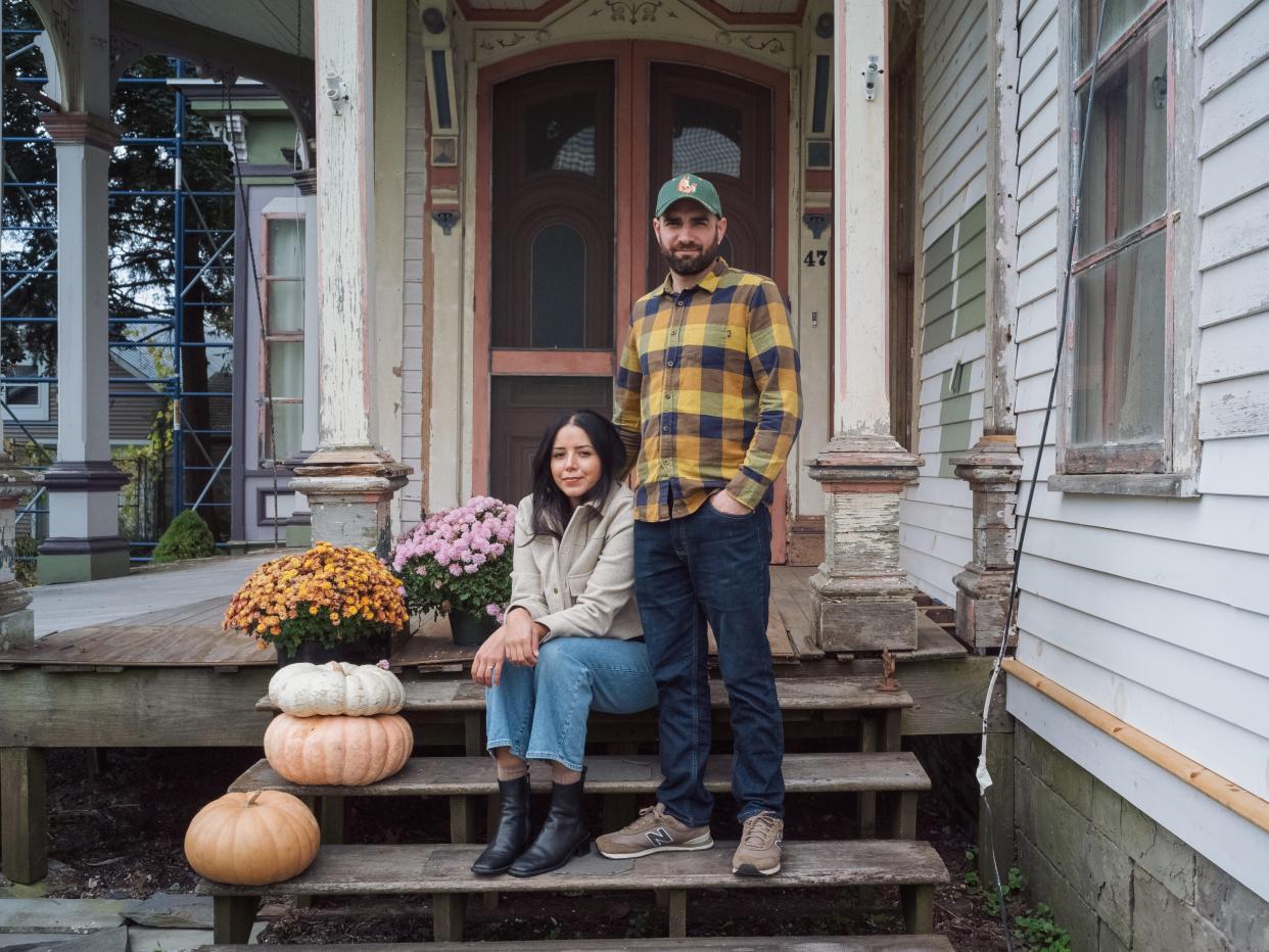 A couple stands on the porch of their dilapidated house with pumpkins and flowers.