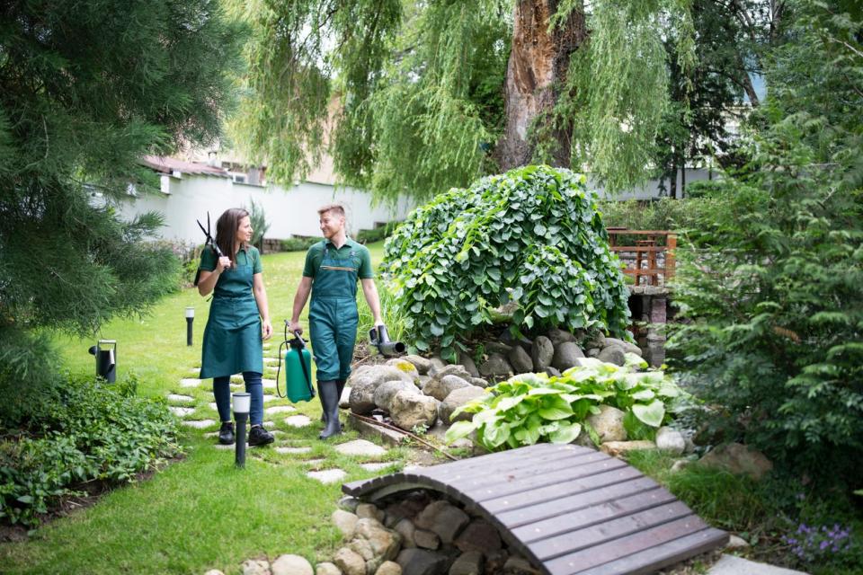 Two workers in green uniforms walk down a garden path holding tools. 