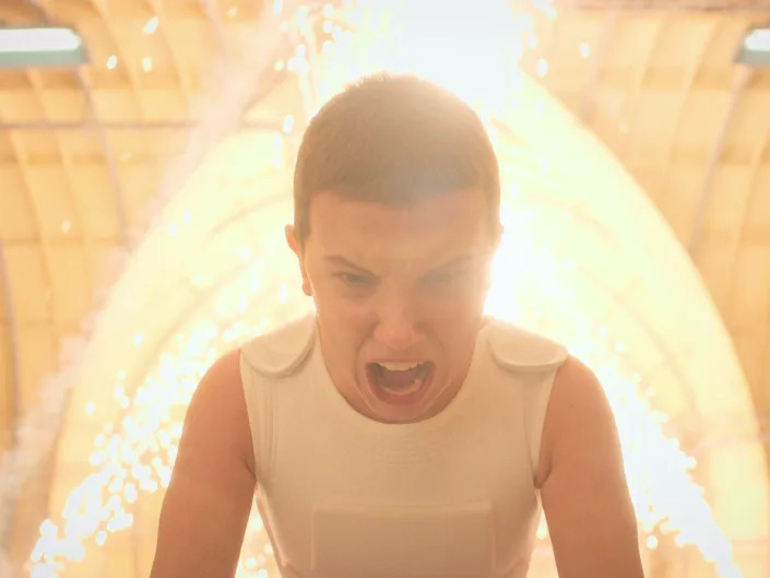 millie bobby brown as eleven in stranger things. her head is shaved and she's wearing a white vest, and as she screams bright yellow sparks arc behind her