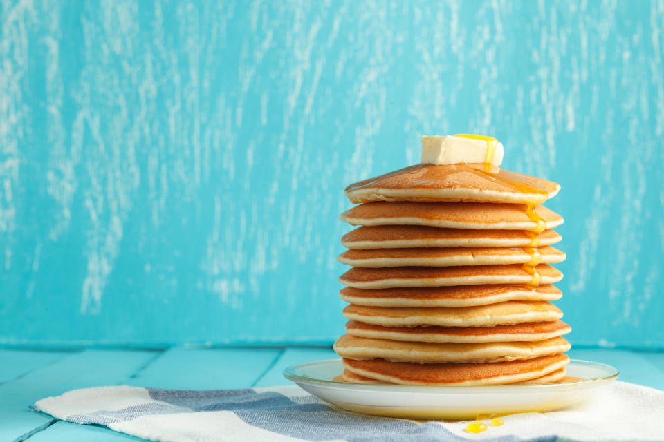 High stack of pancakes on a plate with butter and honey set against a turquoise backdrop.