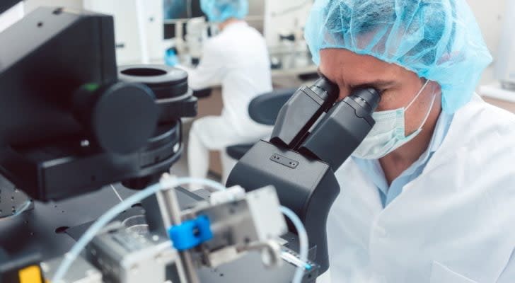 A scientist in medical gear peers through a microscope. IBIO stock