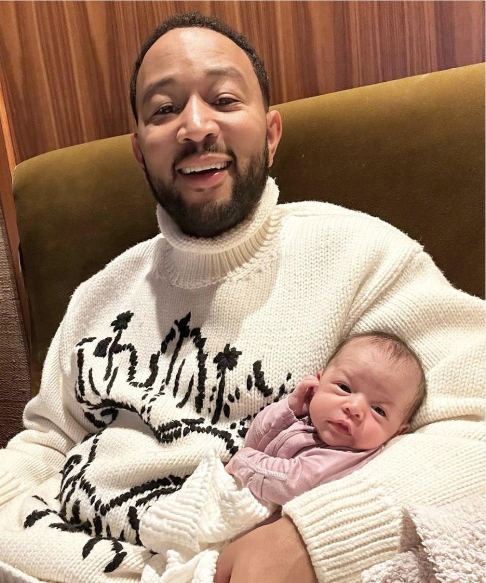 John Legend, in a white turtleneck sweater, smiles at the camera while holding newborn baby Esti, clad in a pink onesie.