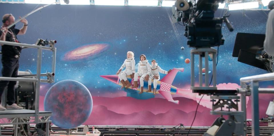 A behind-the-scenes still showing cameras filming the cast of "Barbie" while they ride on top of a rocket on a space-themed set.