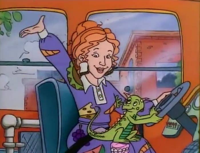 Ms. Frizzle, from "The Magic School Bus," drives the bus while holding a chameleon wearing a small lab coat, with one hand raised enthusiastically