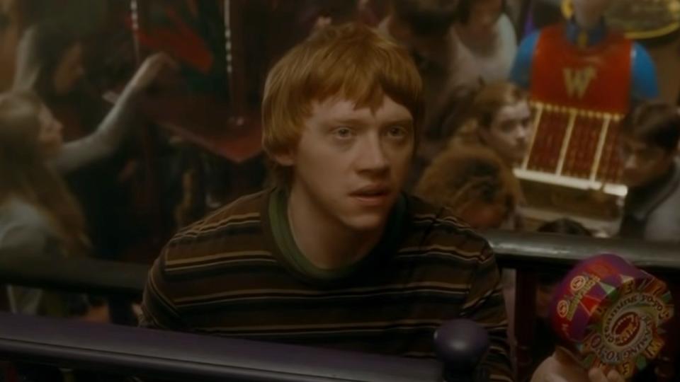 Rupert Grint as Ron Weasley holding a box of candy and scowling in Harry Potter and the Half-Blood Prince