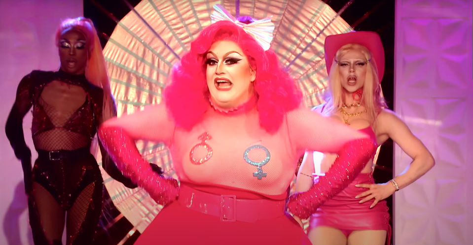 From left to right: Drag performers in extravagant outfits. Center performer wears a pink dress with gender symbols. Scene suggests an LGBT-themed performance