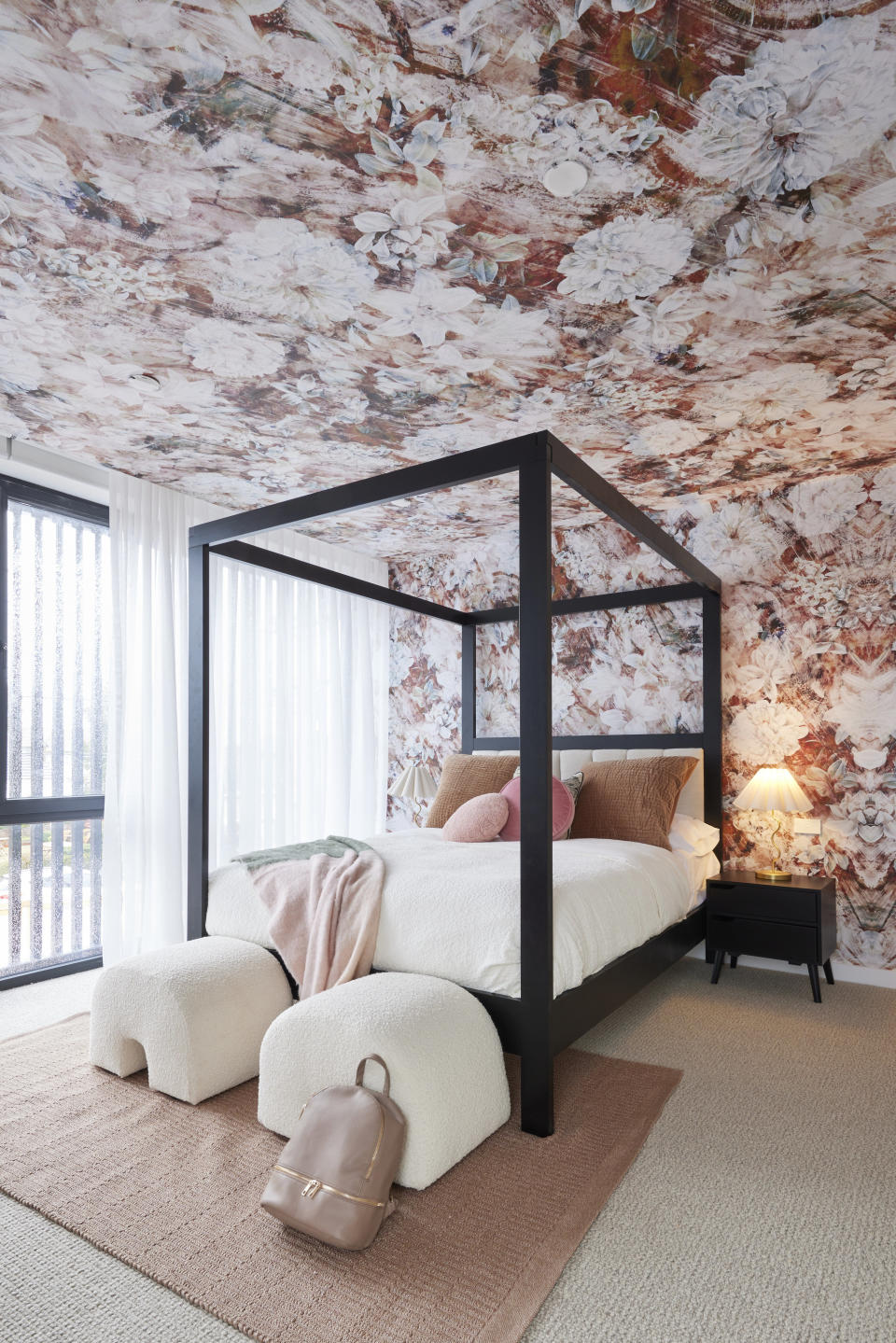 A wide shot of a bedroom with a black four-poster bed and floral wallpaper on the back wall and ceiling. 