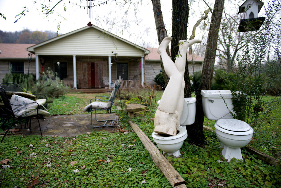 Mannequin legs sticking out of a toilet in a cluttered yard with a house in the background