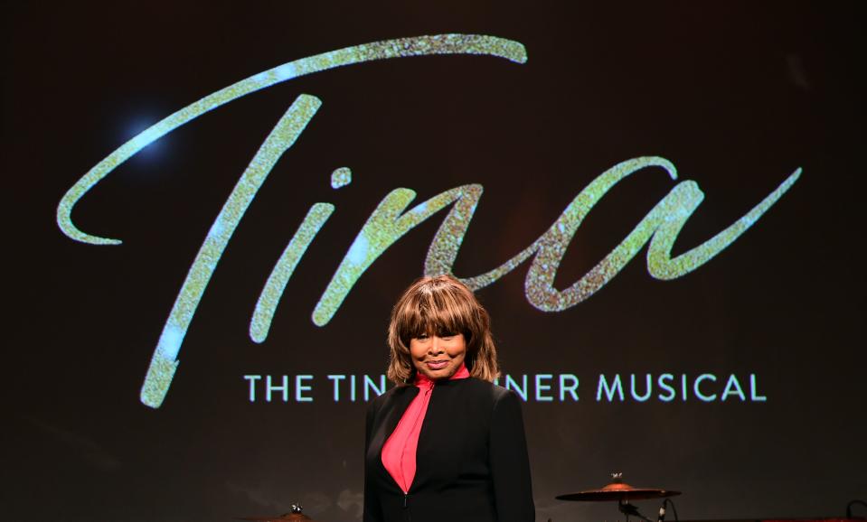 Tina Turner standing in front of a billboard for "Tina: The Tina Turner Musical" while wearing a black blazer and light pink shirt.