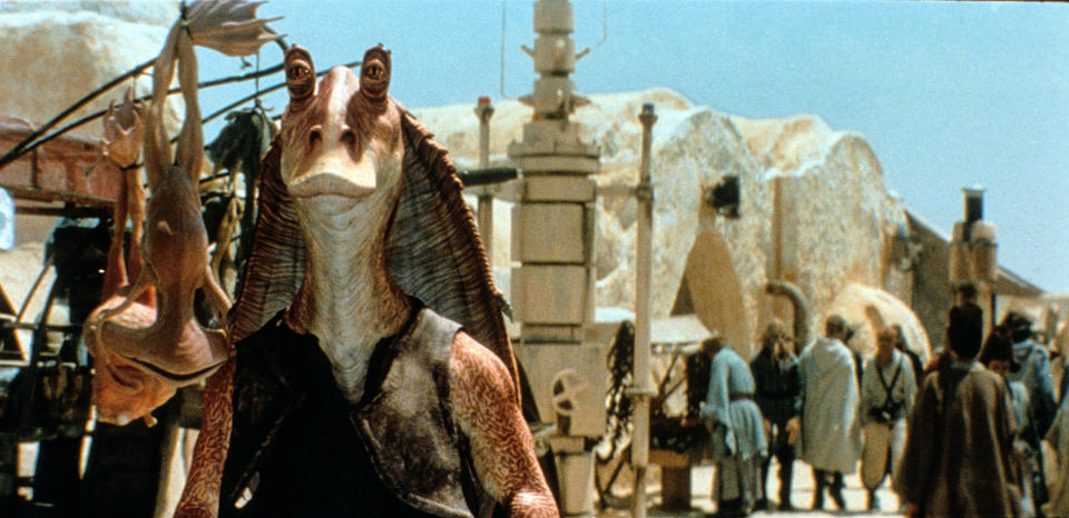 Jar Jar Binks, a character from Star Wars, stands in an outdoor market. There are creatures behind him and people in the background