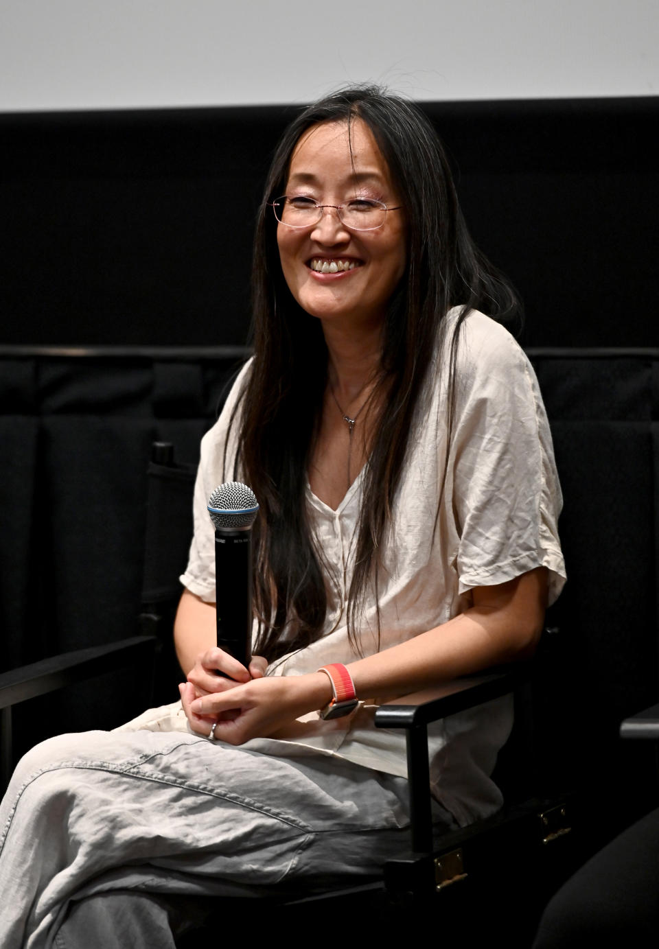 Jennifer Yuh Nelson in a light, casual outfit, holding a microphone while seated on stage at a media event