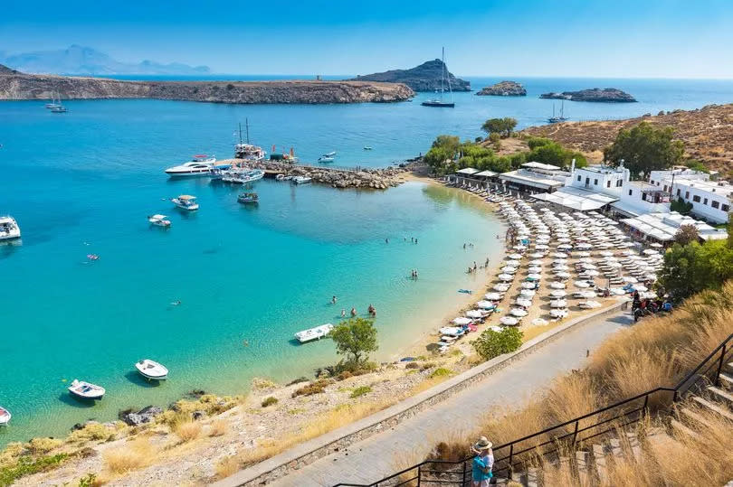 A beach in Greece with parsols and boats in the harbour.