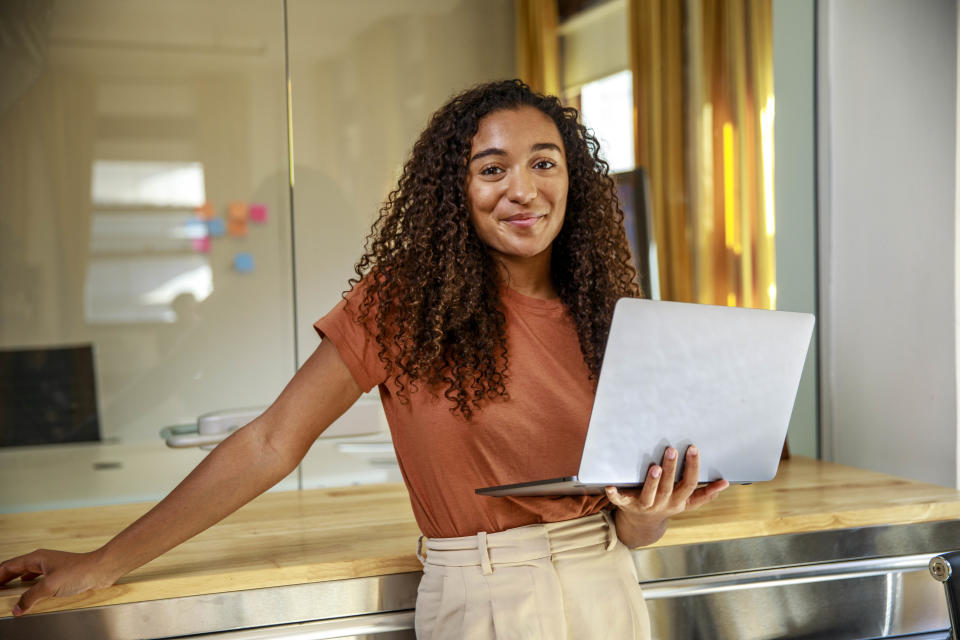 A person stands in a modern office, smiling and holding a laptop