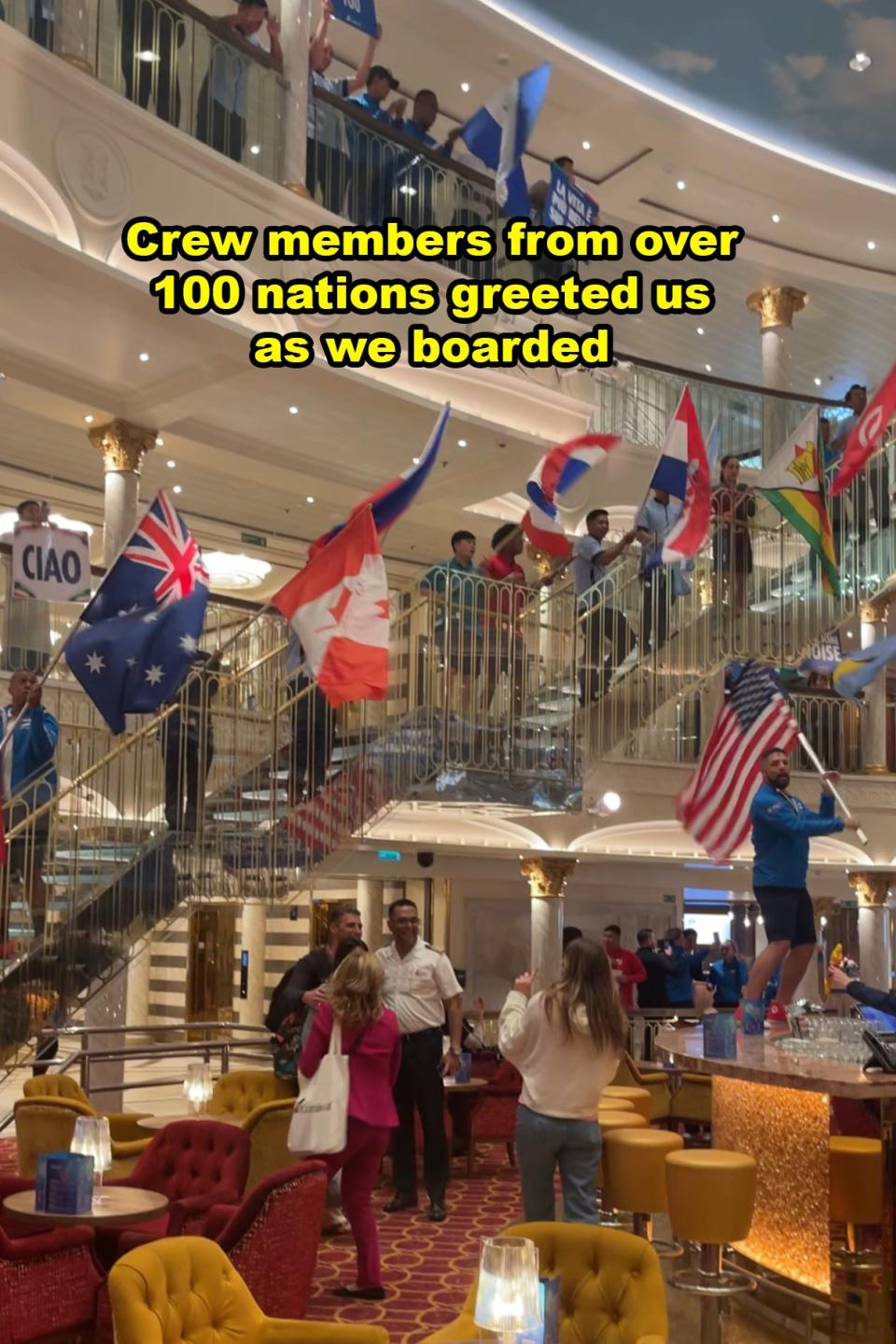 Cruise ship atrium with crew members holding various national flags above welcoming guests