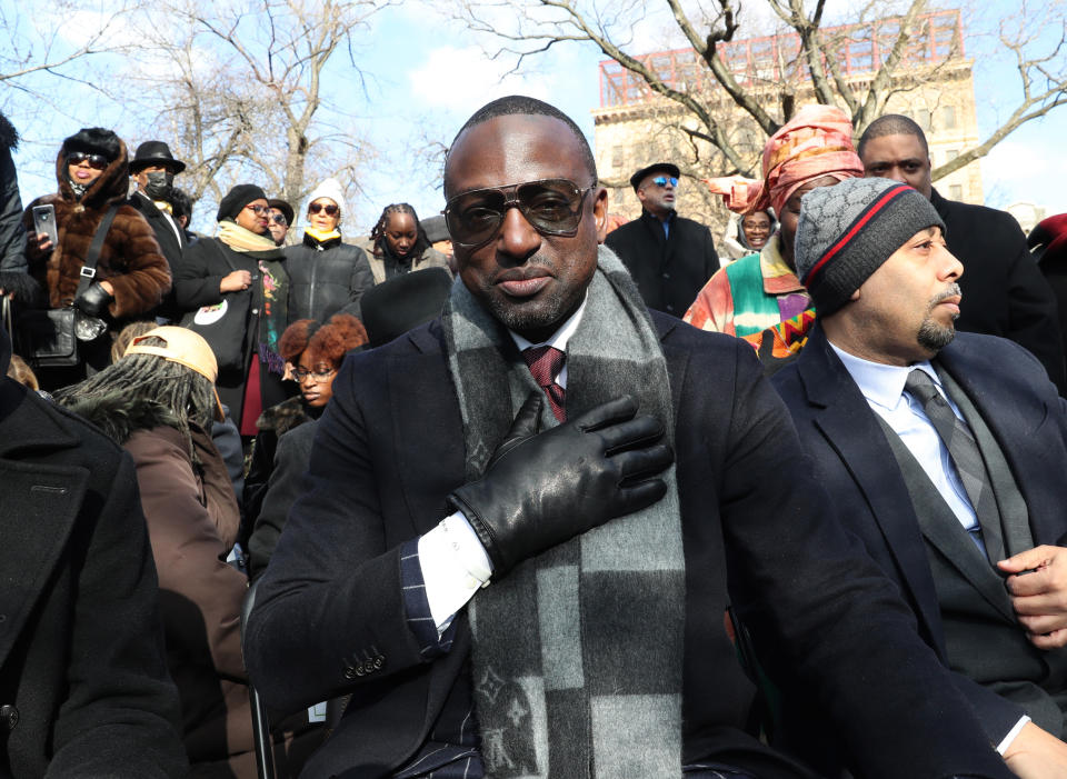 Yusef Salaam, hand on heart, poses in a crowd in Central Park on a cold day, wearing sunglasses, black gloves, a black wool coat and a gray and black checkered scarf.