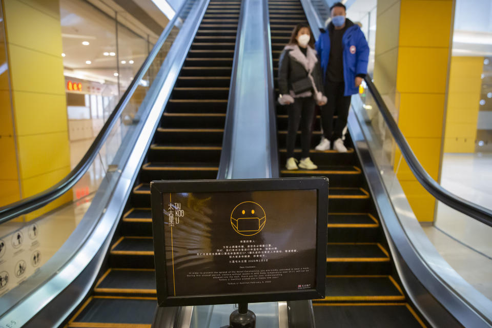 A sign reminds customers to wear face masks as a couple rides an escalator at a shopping center in Beijing, Friday, Feb. 14, 2020. China on Friday reported another sharp rise in the number of people infected with a new virus, as the death toll neared 1,400. (AP Photo/Mark Schiefelbein)