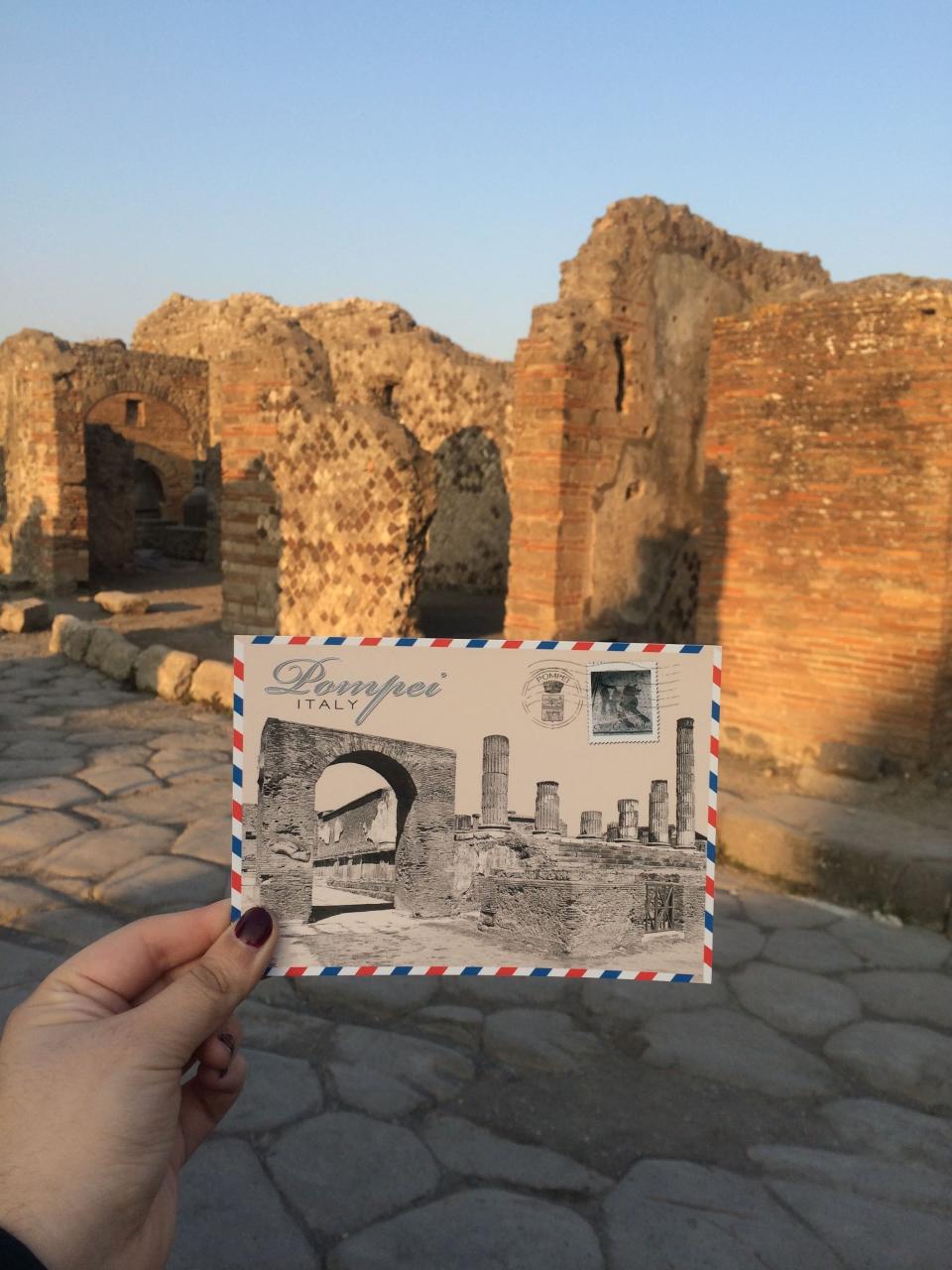 A hand holding up a Pompeii postcard in front of the actual ruins