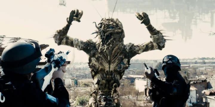 An alien holds up his hands with police aiming their guns at him in "District 9."