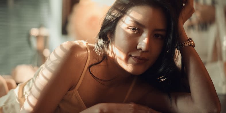 a young woman in a sunny bedroom looking at the camera whilst leaning her head on her hand