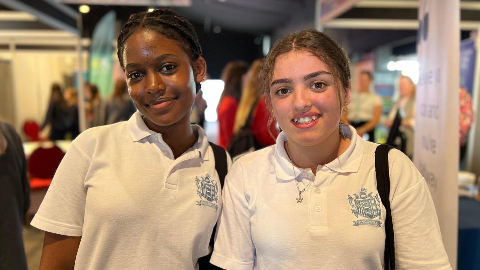 Tiara and Cristina smile at the camera as other students move around exhibits behind them