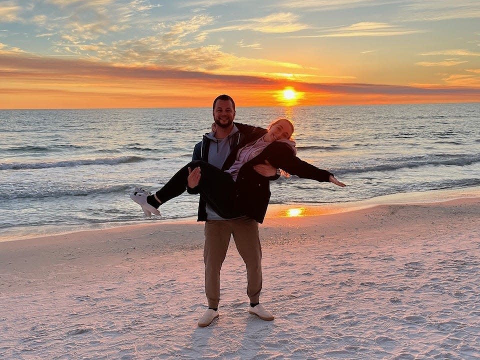 A man jokingly carrying a woman on a beach with a sunset behind.
