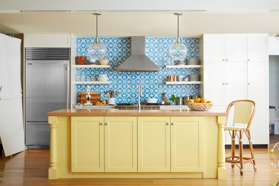 kitchen with yellow painted island and blue and white tile backsplash that extends to the ceiling behind open shelves and stainless steel hood
