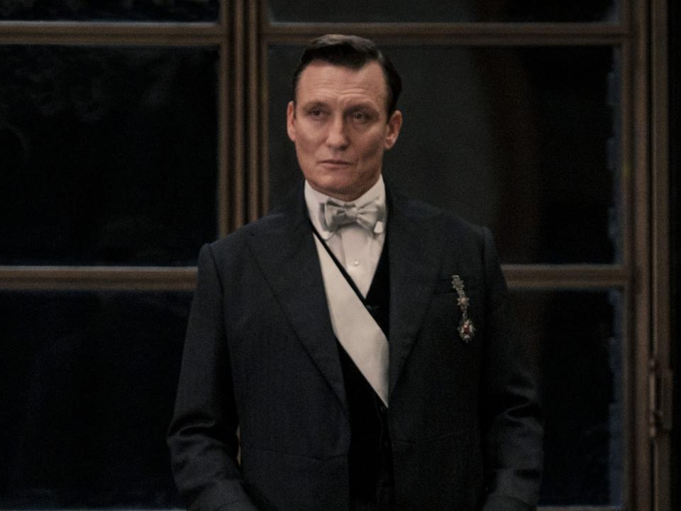 oliver masucci as anton vogel, standing up behind a table in fantastic beasts the secrets of dumbledore