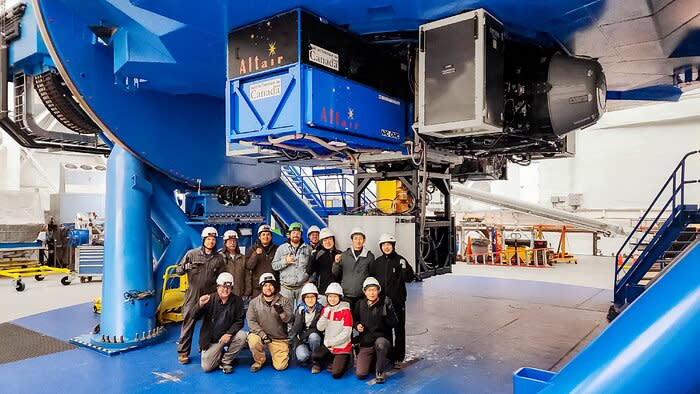 a group of people wearing hard hats pose beneath a portion of a large blue industrial machinery.