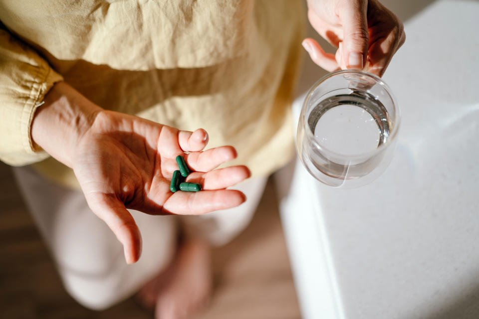 A person holds a green capsule in one hand and a glass of water in the other, possibly preparing to take a supplement or medication.